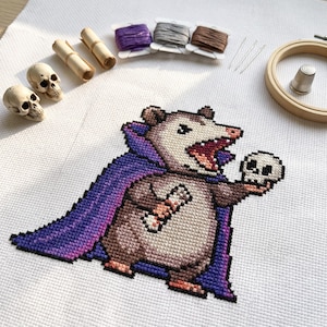 May include: A cross-stitch pattern on white fabric featuring a brown opossum wearing a purple cape, holding a small skull. Accessories include two miniature plastic skulls, three spools of embroidery thread, needles, and a wooden hoop.