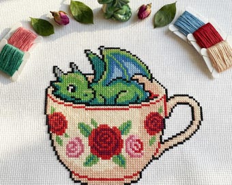 May include: Cross-stitch design of a green dragon in a teacup decorated with red and pink roses. The dragon has blue wings. The image is surrounded by embroidery floss, leaves, and a small ceramic dragon figurine.