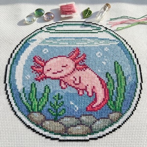May include: A cross-stitch design featuring a pink axolotl swimming in a blue fishbowl. The axolotl has a smiling face and red gills. Green plants and rocks are at the bottom of the bowl. Additional craft supplies are in the background.