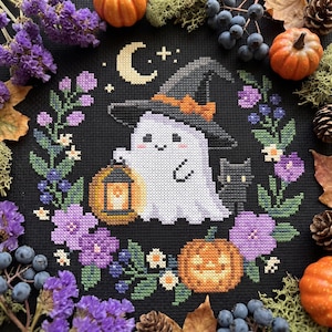 May include: A cross-stitch embroidery on black fabric featuring a white ghost wearing a black witch hat, holding a yellow lantern, and standing next to a black cat. The design is surrounded by a wreath of purple flowers, green leaves, and a pumpkin.