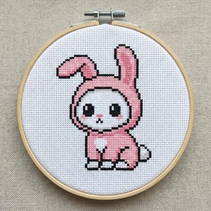 Può includere: Ricamo a punto croce di un simpatico coniglietto con un costume da coniglietto rosa. Il coniglietto ha grandi occhi neri e un piccolo naso rosa. Il ricamo è inserito in un telaio di legno, ideale per l'arredamento murale.