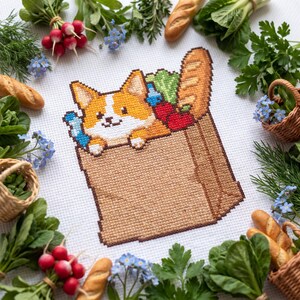 Può includere: Un ricamo a punto croce su tessuto bianco raffigurante un cane Corgi color marrone chiaro e bianco che spunta da un sacchetto della spesa di carta marrone pieno di una baguette, ravanelli rossi e verdure a foglia verde, circondato da fiori.