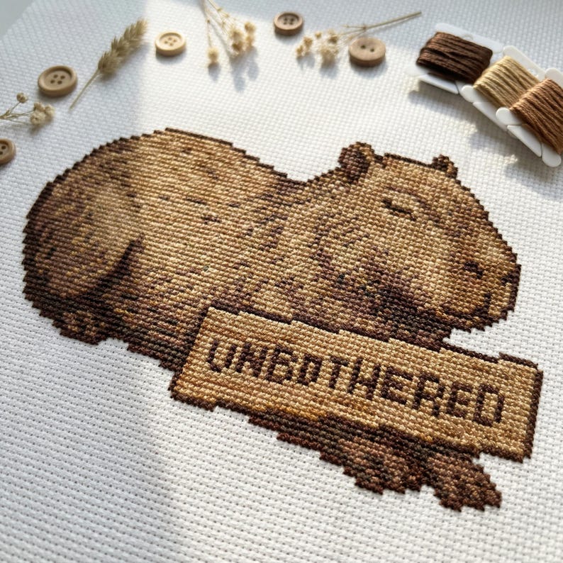 May include: Cross-stitch of a brown capybara with the word "UNBOTHERED" on a sign. The capybara is lying down, stitched on white fabric. Wooden buttons, dried flowers, and spools of thread are arranged around the cross-stitch.