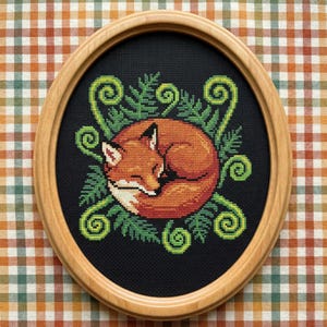 May include: Cross-stitch artwork of a sleeping fox, framed in an oval wooden frame. The fox is orange and white, curled up in a ball, surrounded by green ferns and swirls on a black background. The frame is light brown.