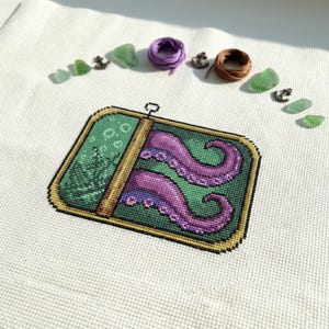 Può includere: Ricamo a punto croce raffigurante una scatola di sardine verde e oro con tentacoli di polpo viola. L'opera è su tessuto bianco, con vetro marino verde, mini ancore e rocchetti di filo viola e marrone.