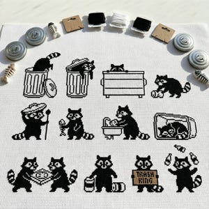 Può includere: Un disegno a punto croce con procioni in bianco e nero in varie pose, tra cui nei bidoni della spazzatura e con pesci. La frase "Trash King" è visualizzata su un cartello. Il disegno è circondato da materiali di cucito ed elementi decorativi.