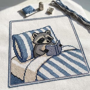 Op de afbeelding: Kruissteekborduurwerk met een wasbeer die in bed leest, met blauw-wit gestreepte beddengoed. Het ontwerp omvat een blauwe rand, een spoel blauw garen, een vingerhoed en een klein boek. Het kleurenpalet is voornamelijk blauw, grijs en wit.