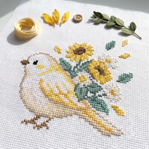 May include: A cross-stitch embroidery on white fabric featuring a light brown bird with yellow accents, surrounded by yellow sunflowers, white daisies, and green leaves. The composition includes a small wooden button, yellow thread, and a green sprig.