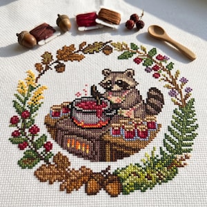 Op de afbeelding: Een kruissteekontwerp met een wasbeer die geniet van een warm drankje, omringd door een krans van herfstbladeren, bessen en eikels. De wasbeer draagt een sjaal. Houten lepel, garenklosjes en eikels zijn rond het ontwerp geplaatst.