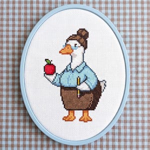 Pode incluir: Bordado em ponto cruz de um ganso usando óculos, camisa azul e saia marrom, segurando uma maçã vermelha e um lápis. A obra de arte é emoldurada em um aro oval azul claro, em um fundo quadriculado.
