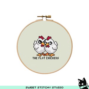 May include: A white embroidery hoop with a cross-stitch design of two chickens with red combs and the text "The Plot Chickens".