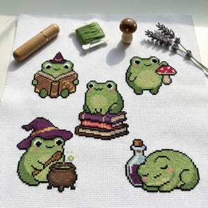 Könnte beinhalten: Kreuzstichmuster mit grünen Fröschen in verschiedenen Szenen auf weißem Stoff. Die Frösche sind beim Lesen, mit einem Pilz und beim Brauen eines Zaubertranks dargestellt. Dazu kommen ein Nadelhalter aus Holz, grünes Garn und ein kleiner Holzpilz.