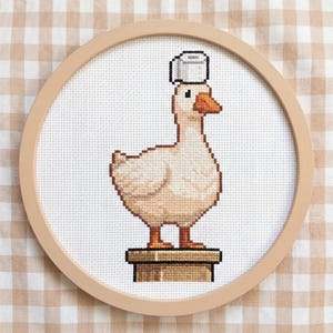 May include: Cross-stitch embroidery of a goose wearing a toilet paper roll on its head, set in a wooden hoop. The goose is beige with an orange beak and feet, standing on a brown platform. The background is white.