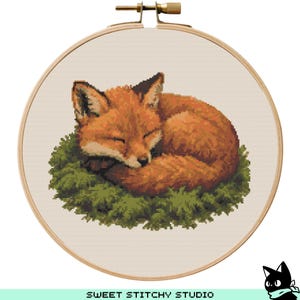 May include: A cross-stitch embroidery hoop featuring a sleeping fox with orange fur, nestled on a bed of green foliage. The fox is curled up, eyes closed, and the design is set against a cream-colored fabric. The text "SWEET STITCHY STUDIO" is at the bottom.