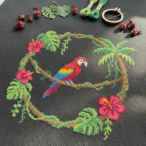 May include: Cross-stitch embroidery featuring a vibrant parrot on a branch, encircled by a wreath of tropical flowers and foliage. The parrot displays red, yellow, blue, and green plumage. The embroidery is set against a dark grey fabric.