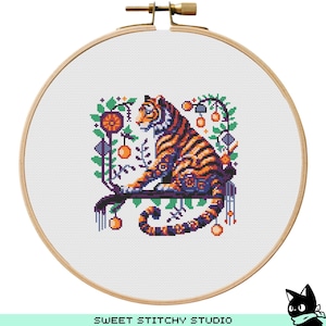May include: A cross-stitch embroidery hoop featuring a tiger design. The tiger has orange and black stripes and is perched on a branch with decorative elements. The background is white, and the hoop is made of wood. The text "SWEET STITCHY STUDIO" is at the bottom.