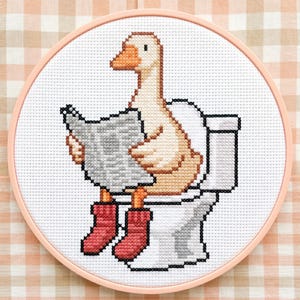 Könnte beinhalten: Kreuzstich-Stickerei einer Gans, die auf einer Toilette sitzt, eine Zeitung liest und rote Stiefel trägt. Die Gans ist in Beige-, Orange- und Weißtönen gehalten. Die Stickerei befindet sich in einem rosa Reifen vor einem karierten Hintergrund.