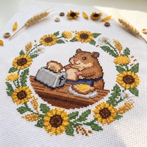 May include: A cross-stitch embroidery on white fabric featuring a brown hamster sitting at a wooden table with a silver toaster and a slice of toast. The hamster is surrounded by a circular wreath of yellow sunflowers and green leaves.