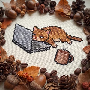 May include: A cross-stitch embroidery on cream fabric featuring a pixelated orange tabby cat lying on a gray laptop computer with a brown mug of steaming coffee nearby, surrounded by a border of dried autumn leaves, acorns, and coffee beans.