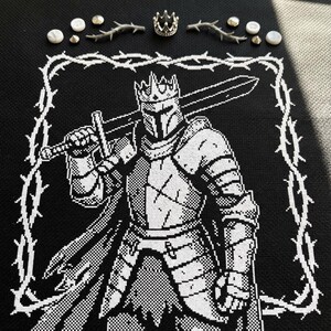 Può includere: Design a punto croce in bianco e nero di un cavaliere in armatura, con una spada e una corona. Il cavaliere è incorniciato da un bordo spinoso bianco. Elementi decorativi: corona d'argento, spine e perle.