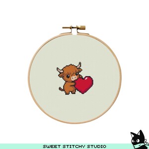 May include: A cross-stitch embroidery hoop featuring a brown Highland cow holding a red heart. The cow has small horns and a fluffy coat. The background is a light beige fabric. The wooden hoop is secured with a brass clasp. The text "SWEET STITCHY STUDIO" is at the bottom.