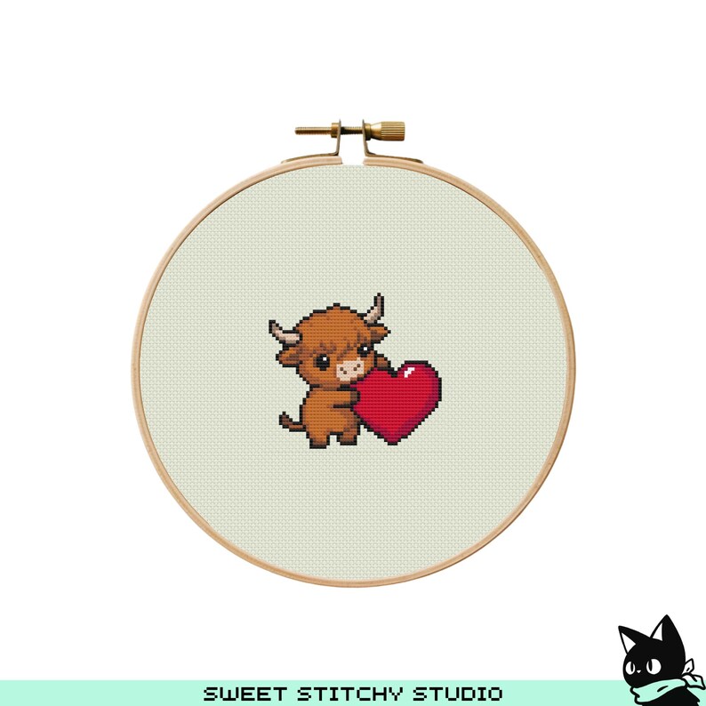 May include: A cross-stitch embroidery hoop featuring a brown Highland cow holding a red heart. The cow has small horns and a fluffy coat. The background is a light beige fabric. The wooden hoop is secured with a brass clasp. The text "SWEET STITCHY STUDIO" is at the bottom.
