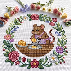 May include: A cross-stitch embroidery on white fabric featuring a brown mouse wearing a purple apron, grating yellow cheese on a wooden table. The mouse is centered within a circular wreath of colorful flowers, including roses, daisies, and bluebells.