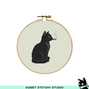 Puede incluir: Bordado de punto de cruz en un aro de madera que muestra un gato negro con ojos verdes fumando un cigarrillo. El gato está sentado y el humo se eleva del cigarrillo. El fondo es de tela beige claro.