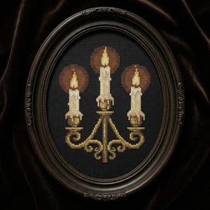 May include: Cross-stitch artwork of three lit candles in a gold candelabra, set against a black background. The candles have yellow flames and are framed in an oval, dark brown frame. The artwork is displayed on a dark brown velvet background.