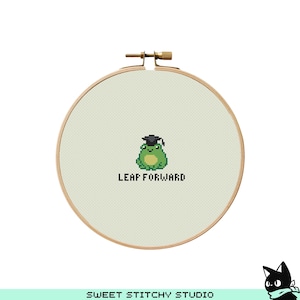 May include: Cross-stitch embroidery hoop featuring a green frog wearing a graduation cap. The text "LEAP FORWARD" is below the frog. The hoop is made of light beige fabric and a wooden frame.