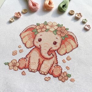 Peut inclure: Point de croix d'un éléphant bébé avec une couronne de fleurs. L'éléphant est dans des tons pêche et rose, avec des yeux bruns. Des fils à broder et des boutons en forme de fleurs entourent le motif, sur fond blanc.