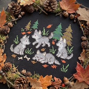 May include: A cross-stitch embroidery on black fabric featuring three gray wolves, green pine trees, and scattered orange autumn leaves. The design includes two sitting wolves, one standing wolf, and one lying wolf, accented with small green grass tufts.