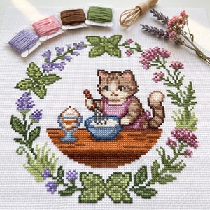 May include: A cross-stitch embroidery on white fabric featuring a brown tabby cat wearing a pink apron, mixing ingredients in a blue bowl on a wooden table. The cat is surrounded by a circular wreath of green leaves, purple flowers, and pink blossoms.