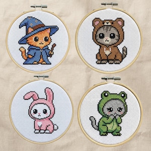 May include: Four cross-stitch hoops featuring cute cat designs. One cat is dressed as a wizard, another as a bear, a third as a bunny, and the last as a frog. Each design is stitched on white fabric and framed in a wooden hoop.