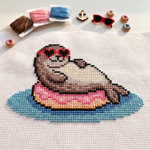 May include: A cross-stitch pattern on white fabric featuring a brown seal wearing red heart-shaped sunglasses while floating on a pink doughnut-shaped pool float in blue water. The scene includes embroidery floss spools, a small anchor, and tiny doughnuts.