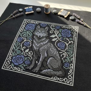 May include: Cross-stitch of a gray wolf with blue eyes, surrounded by blue roses and green foliage on a black background. The wolf is seated, with detailed shading. The design is framed with a white border. Spools of thread and lavender sprigs are arranged around the cross-stitch.