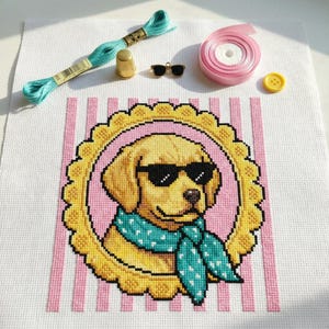 Może przedstawiać: Projekt haftu krzyżykowego przedstawiający złotego retrievera w okularach przeciwsłonecznych i turkusowym szaliku w białe grochy. Pies jest oprawiony w żółte, faliste koło na różowym, pasiastym tle. Dodatkowe materiały rzemieślnicze obejmują turkusową nić, naparstek i różową miarkę.