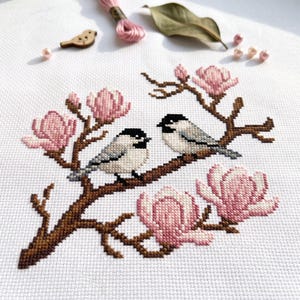 May include: A cross-stitch embroidery on white fabric featuring two small birds perched on a brown branch surrounded by pink magnolia flowers. The design includes a wooden bird-shaped button, pink embroidery floss, and small beads.
