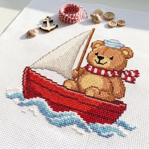 Può includere: Un ricamo a punto croce su tessuto bianco raffigurante un orsacchiotto marrone con un cappello da marinaio blu e una sciarpa a righe rosse e bianche, seduto su una barca a vela rossa con vela bianca su onde blu, con un'ancora di legno.