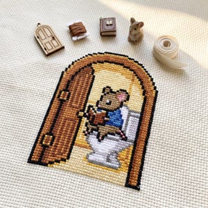 May include: Cross-stitch art featuring a mouse in a blue jacket sitting on a toilet inside a brown door frame. Additional elements include a wooden door, thread, a miniature book, a small mouse figurine, and a ribbon.