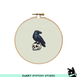 Puede incluir: Un bastidor de bordado de punto de cruz con un cuervo negro posado sobre una calavera blanca. El cuervo y la calavera están pixelados. El fondo es de tela beige claro. El bastidor de madera es visible. El texto "SWEET STITCHY STUDIO" está en la parte inferior.
