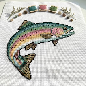 Puede incluir: Un diseño de punto de cruz de una trucha arcoíris en tonos verdes, azules, rosas y amarillos. El pez está sobre un fondo blanco, con tres carretes de hilo y pequeñas piedras. El pez está saltando, con la boca abierta.