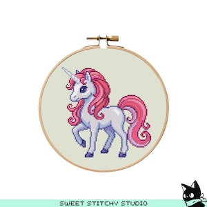 Op de afbeelding: Kruissteekborduurwerk van een eenhoorn in een houten hoepel. De eenhoorn is wit met een roze manen en staart, en een zilveren hoorn. De achtergrond is een lichtgroene stof. De tekst "SWEET STITCHY STUDIO" staat onderaan.