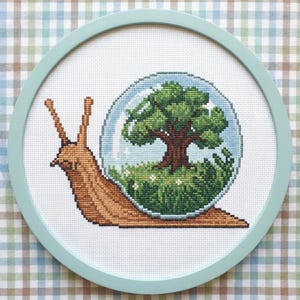 Könnte beinhalten: Kreuzstich-Kunstwerk einer Schnecke mit einer Glaskugel-Schale. In der Kugel befinden sich ein Baum, Gras und Blumen. Die Schnecke ist braun und der Rahmen hellblau. Der Hintergrund hat ein kariertes Muster.