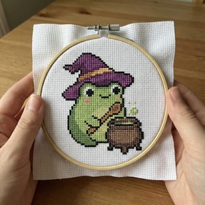 Pode incluir: Bordado em ponto cruz de um sapo verde usando um chapéu de bruxa roxo com uma faixa dourada, mexendo um caldeirão marrom com uma colher de pau. O sapo tem uma expressão sorridente. O bordado está em um bastidor de madeira.