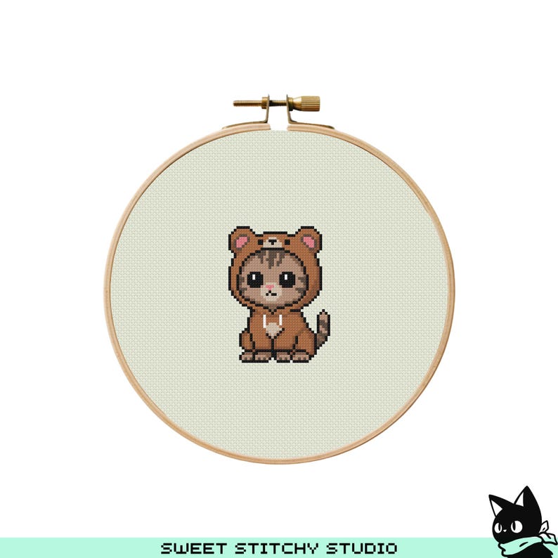 May include: Cross-stitch embroidery of a cat wearing a brown bear costume. The cat is brown and tan with a cute expression. The embroidery is set in a wooden hoop. The text "SWEET STITCHY STUDIO" is at the bottom.