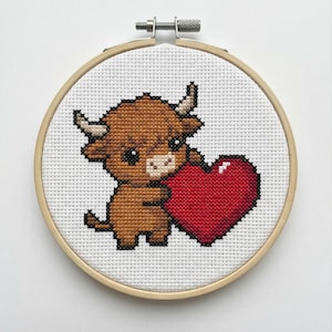 May include: A cross-stitch embroidery hoop featuring a brown Highland cow holding a red heart. The cow has small horns and big eyes. The embroidery is set in a wooden hoop against a white background.