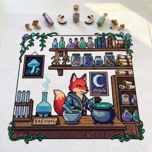 Puede incluir: Arte de punto de cruz que representa un zorro con una túnica de mago, preparando pociones. La escena incluye estantes con tarros, botellas y libros, además de una imagen de hongo enmarcada. La palabra "BREWING" es visible. Setas de madera y botellas decorativas están dispuestas alrededor de la obra.