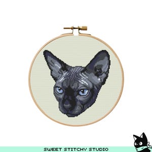 May include: A cross-stitch embroidery hoop showing a black Sphynx cat with blue eyes. The cat's head is centered on a light beige fabric, framed by a wooden hoop. The text "SWEET STITCHY STUDIO" is at the bottom.