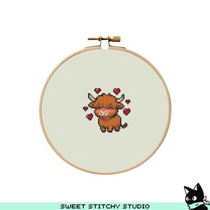 May include: A cross-stitch embroidery hoop featuring a brown Highland cow with a pink blush and surrounded by red hearts. The text "SWEET STITCHY STUDIO" is at the bottom. The hoop is made of wood and has a metal clasp.